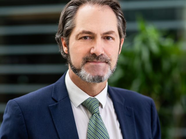 Dr. Andre Bachmann wearing a blue suit jacket and green tie with a white shirt posed in front of a blurred backdrop of a grey wall with a green plant