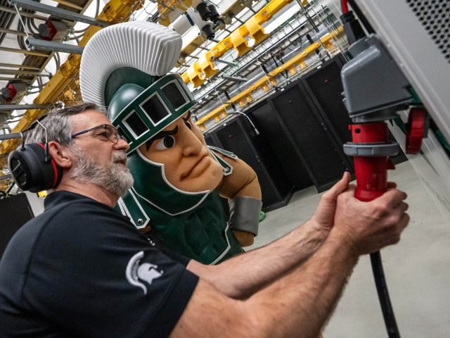 ICER system administrator Kelly Climer shows Sparty how to plug in MSU’s new 2025 supercomputing hardware cluster.